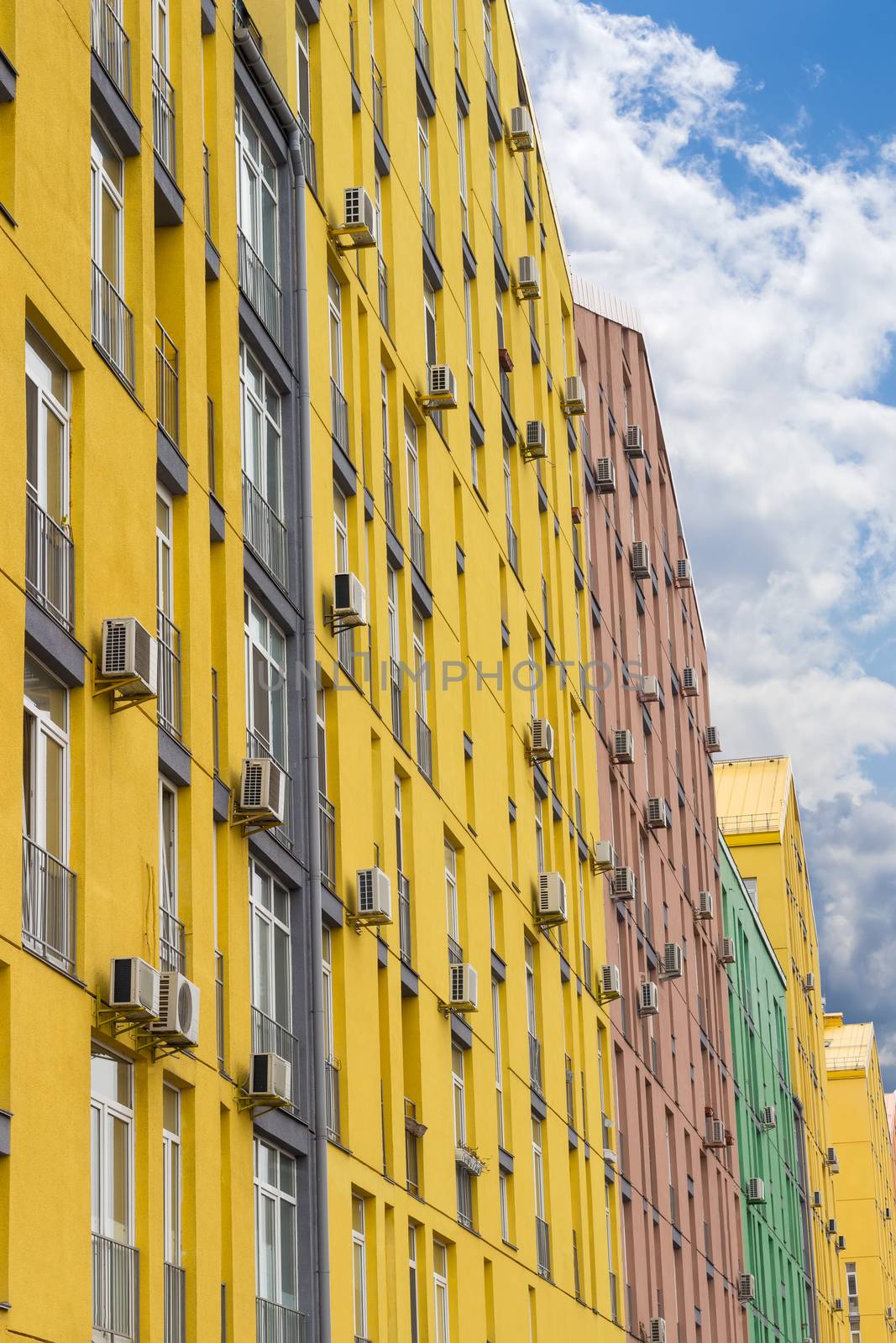 Fragment of a multicolored facade of the modern multi-story apartment complex with the outside parts of a split-type air conditioners against the background of the sky with clouds
