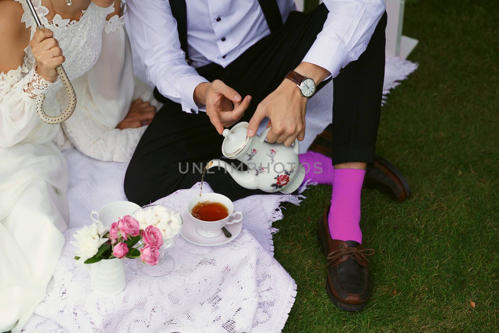Bride and groom drink tea in bright clothes on the green grass