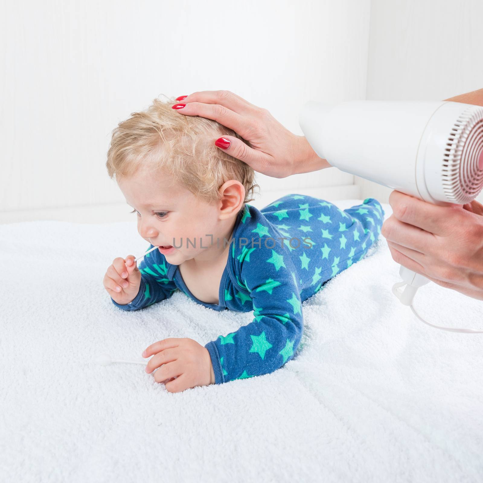 Mom dries baby boy hair with a hair dries, while the infant toys lying with cotton swab.