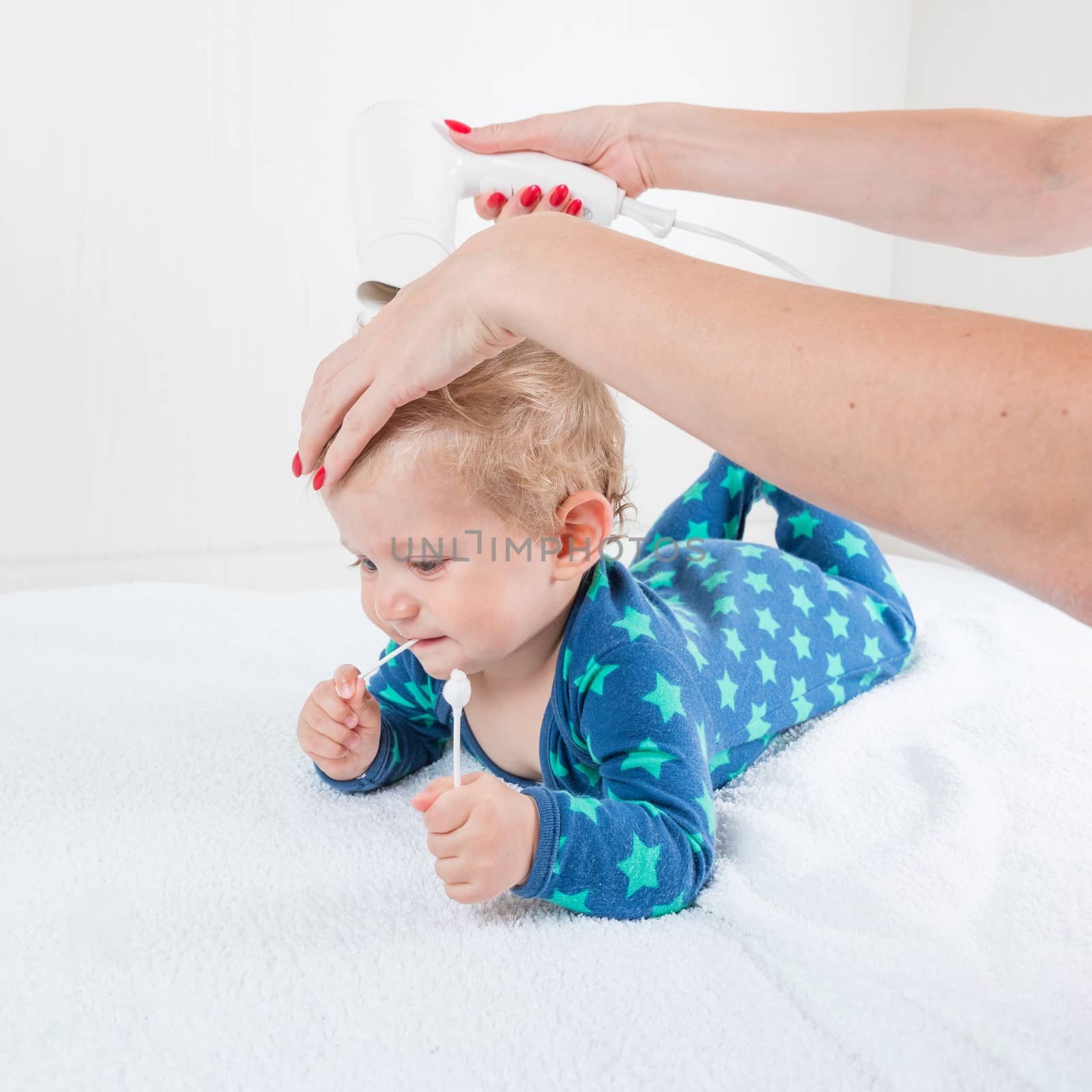 Mom dries baby boy hair with a hair dries, while the infant toys lying with cotton swab.