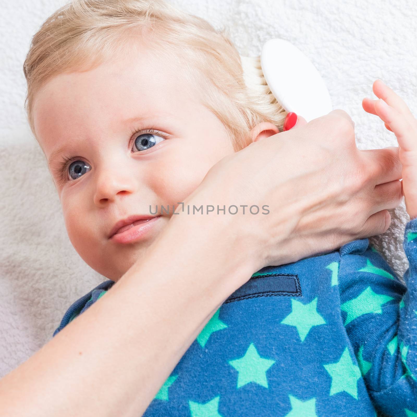 Mom combing the baby with blonde hair, while the toddler is lying.