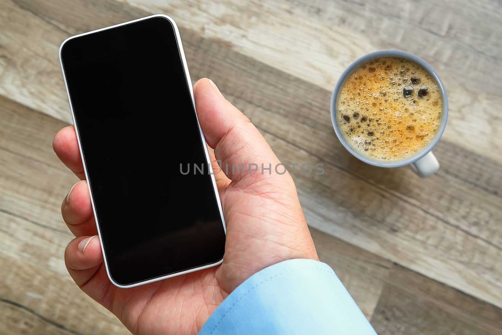 Smartphone in hand at work with a blank area to insert a custom image. Cup of tea and wooden floor on background