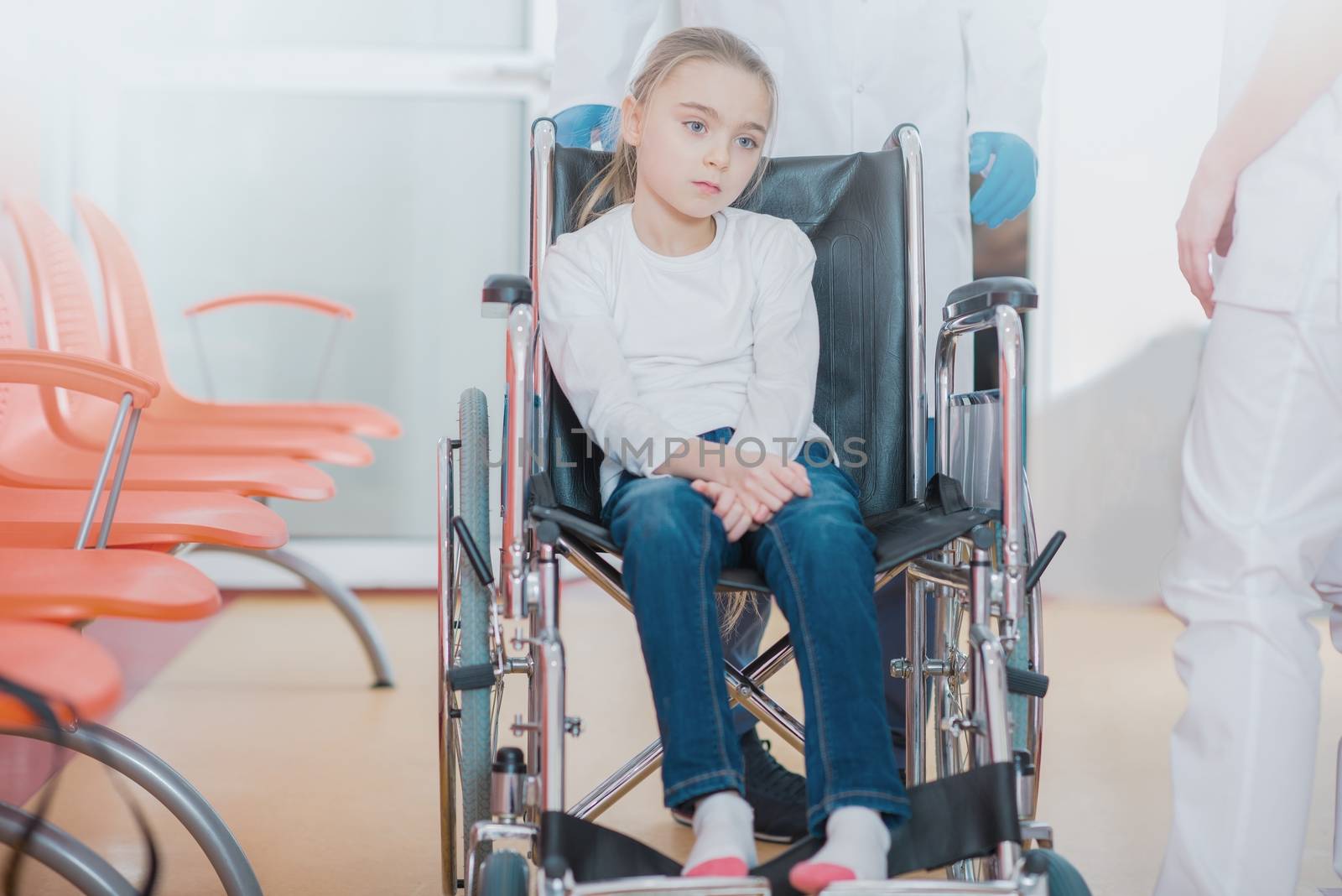Wheelchair Disability in Children. Mobility Impairments. Young Caucasian Girl on the Wheelchair in the Hospital Hall with Physicians in the Background.