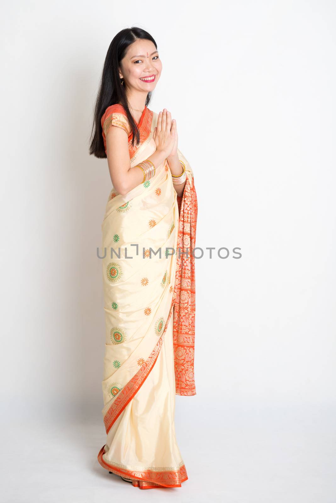 Full length mixed race Indian Chinese girl with sari dress in greeting gesture, standing on plain background.