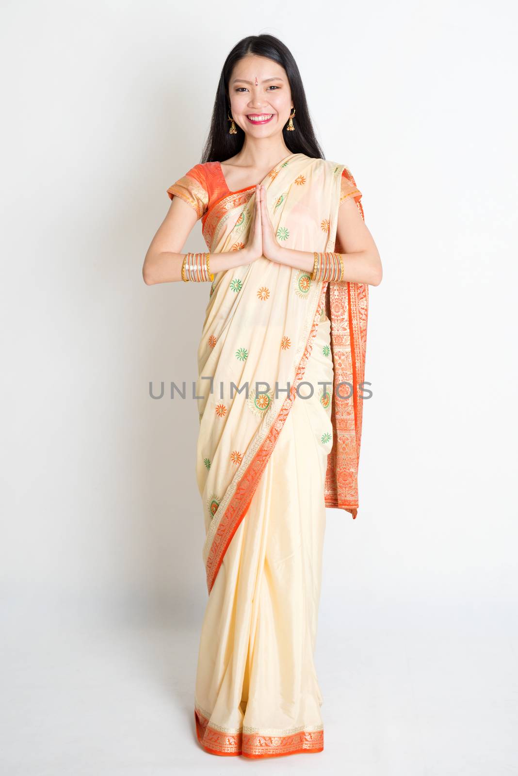 Full length mixed race Indian Chinese female with sari dress in greeting gesture, standing on plain background.