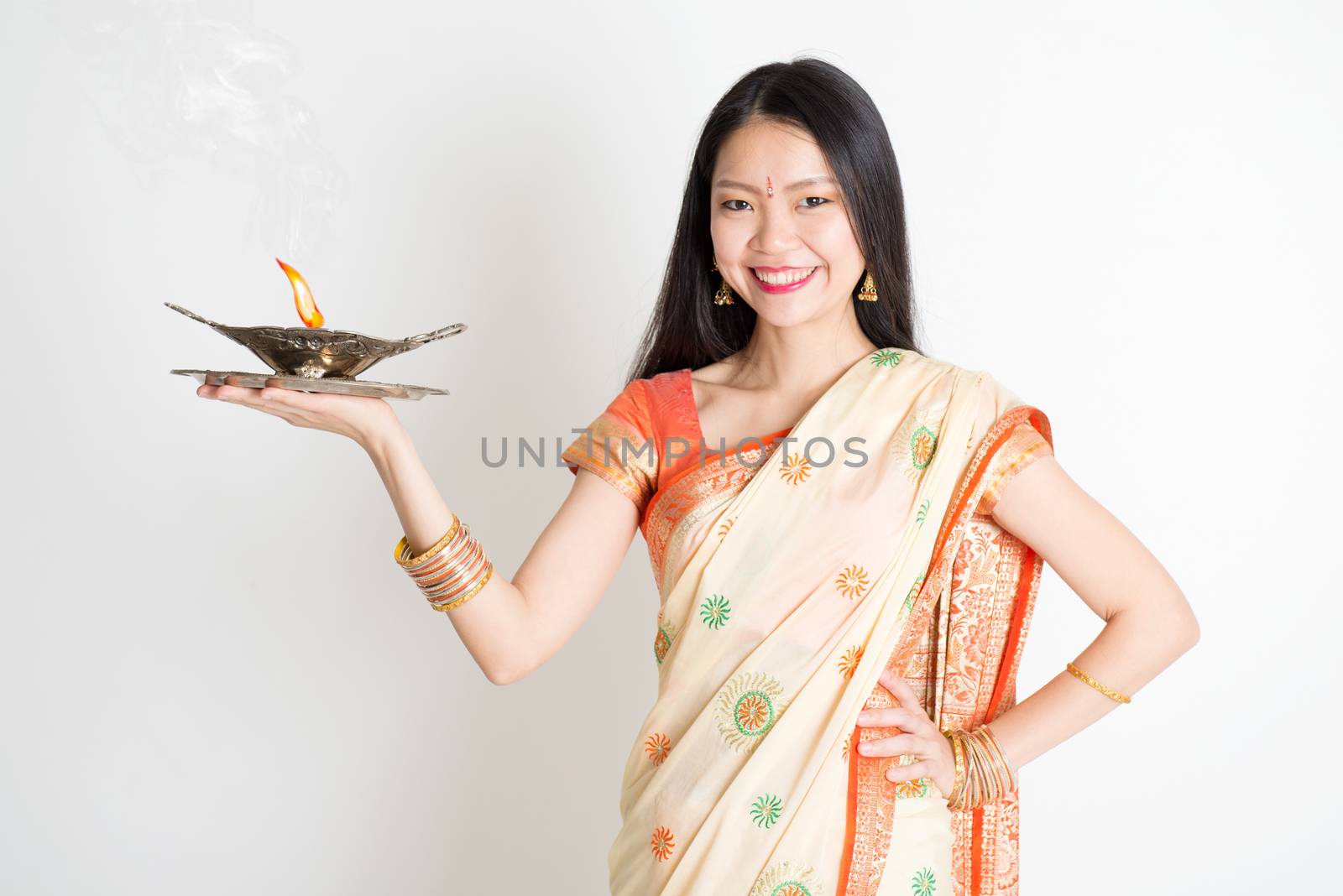 Portrait of young mixed race Indian Chinese female in traditional sari dress, holding diya oil lamp light, on plain background.