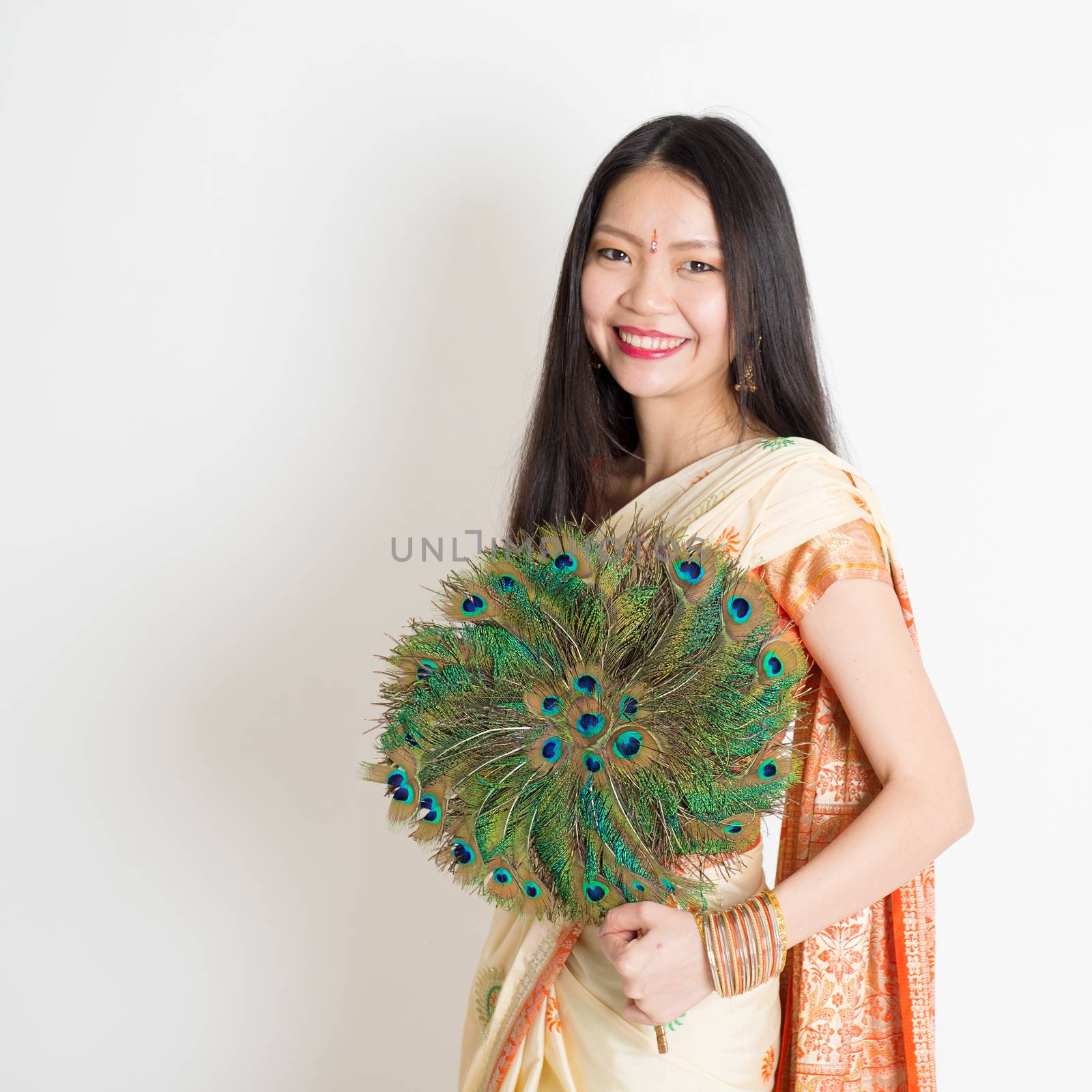 Portrait of young mixed race Indian Chinese female in traditional sari dress, holding peacock feathers fan and looking at camera, on plain background.