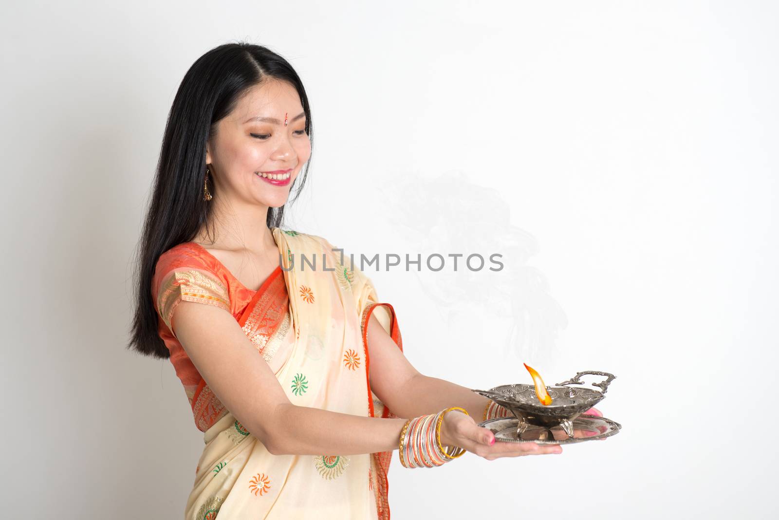 Portrait of young mixed race Indian Chinese female in traditional sari dress, holding oil lamp, on plain background.