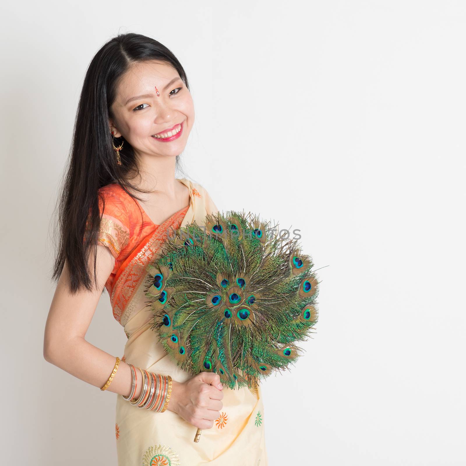 Portrait of young mixed race Indian Chinese woman in traditional sari dress, holding peacock feathers fan and looking at camera, on plain background.