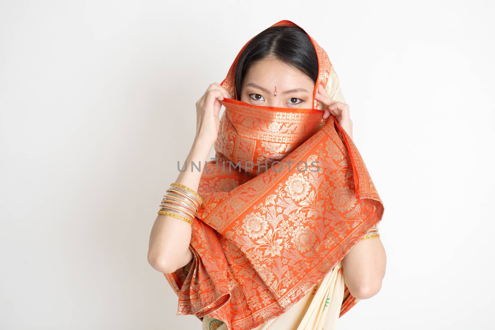 Portrait of secrecy mixed race Indian Chinese woman in traditional sari dress, covering face and looking at camera, on plain background.