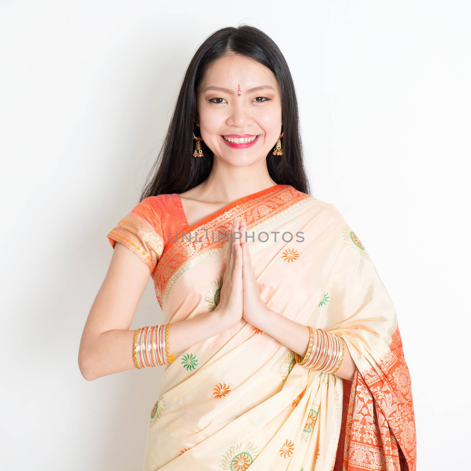 Portrait of mixed race Indian Chinese girl with traditional sari dress in greeting gesture, standing on plain background.