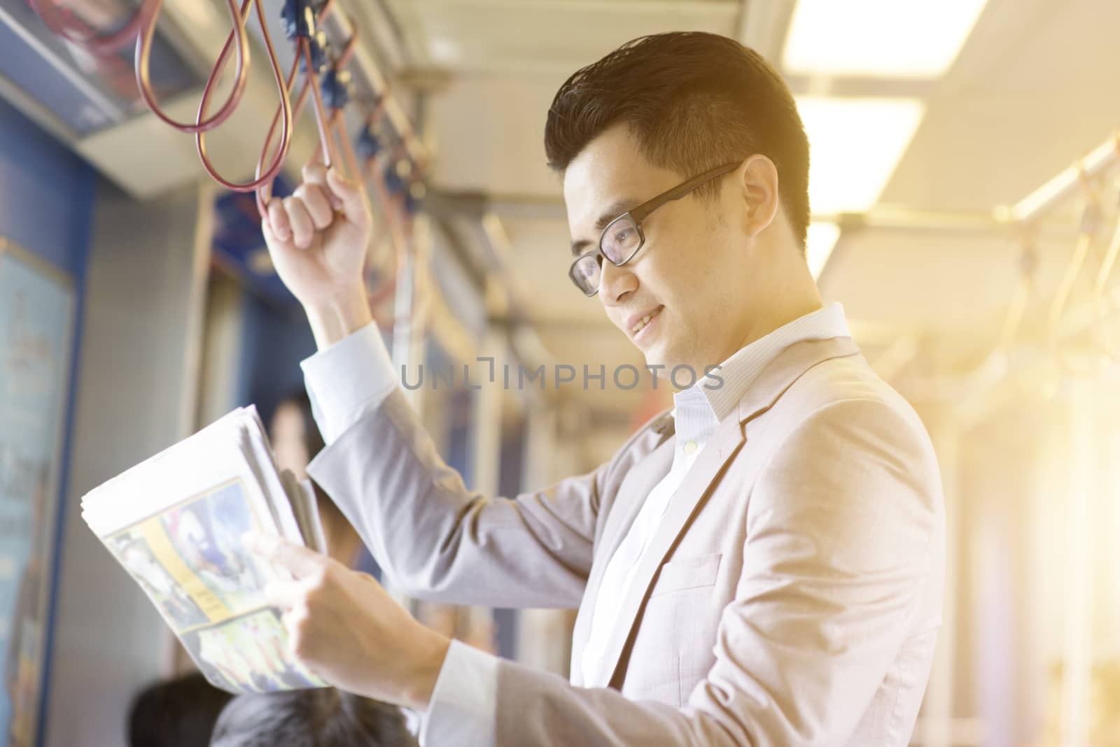 Asian Chinese businessman taking ride to work in morning, standing inside public transport and reading newspaper.