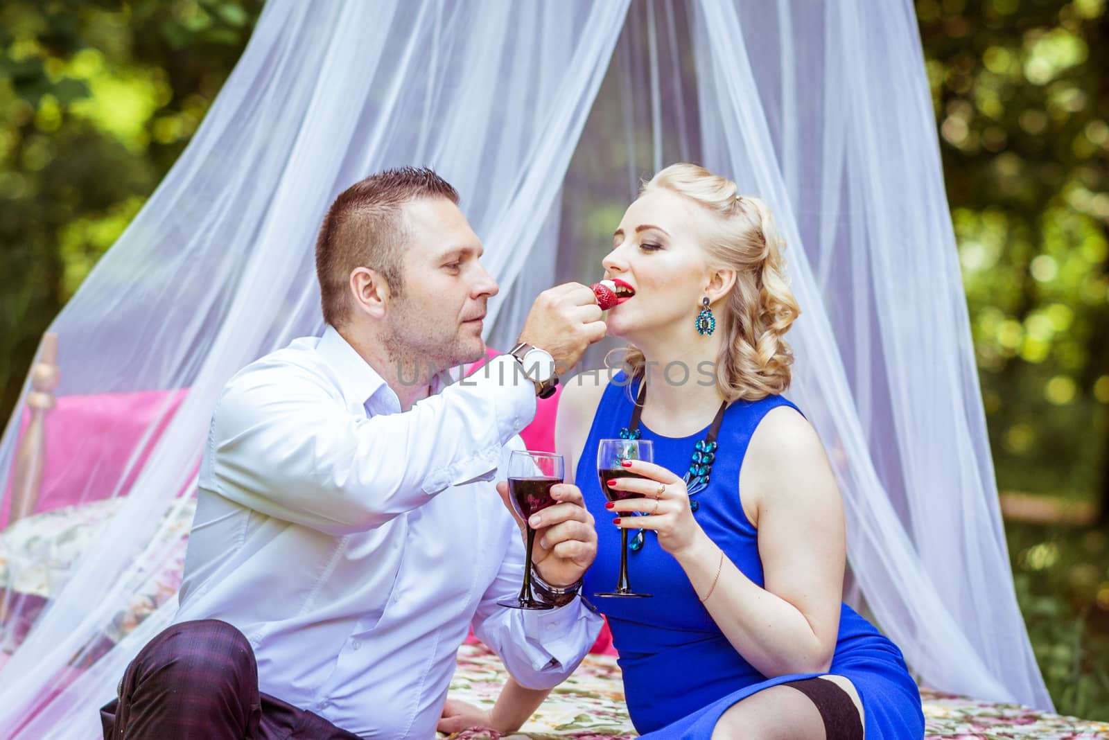 Man and woman sitting on the bed in the lawn with glasses of wine and woman eating strawberries from a man's hand in Lviv, Ukraine.