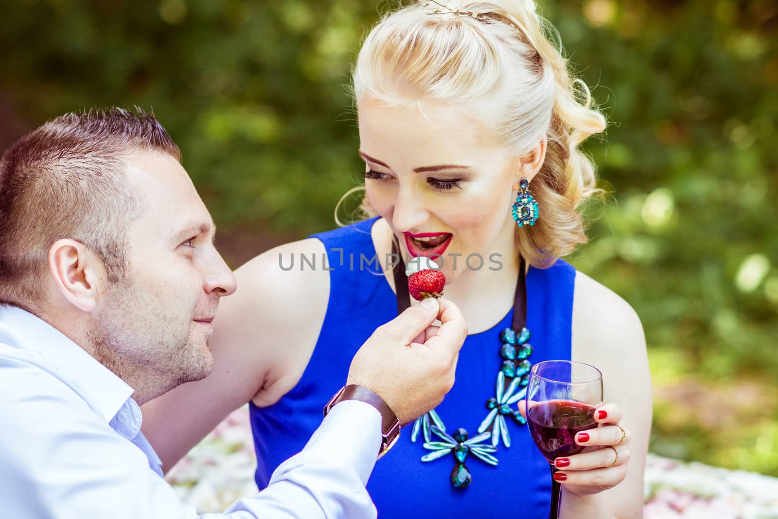 Man and woman sitting on the bed in the lawn with glasses of wine and woman eating strawberries from a man's hand in Lviv, Ukraine.