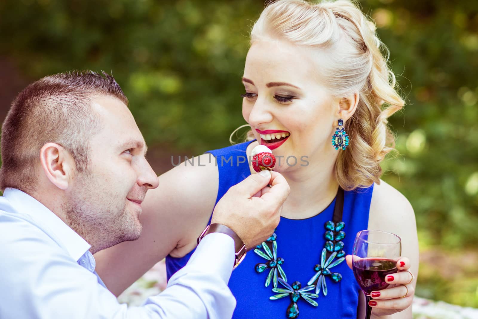 Man and woman sitting on the bed in the lawn with glasses of wine and woman eating strawberries from a man's hand in Lviv, Ukraine.