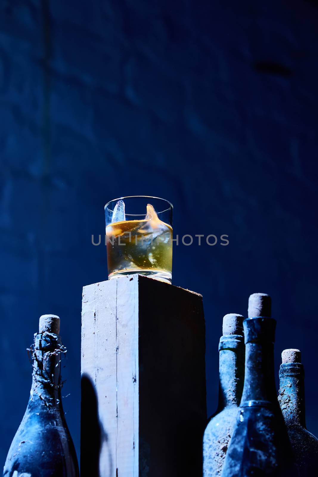 A glass of whiskey with ice on old wooden bar around the old bottles. Shallow dof. The blue tones