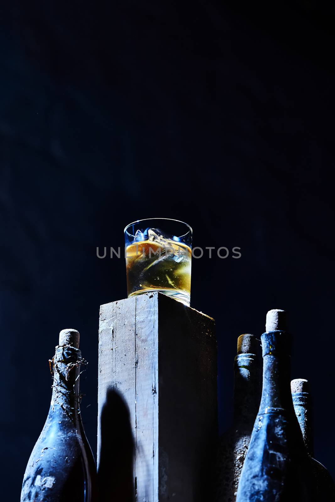 A glass of whiskey with ice on old wooden bar around the old bottles. Shallow dof. The blue tones