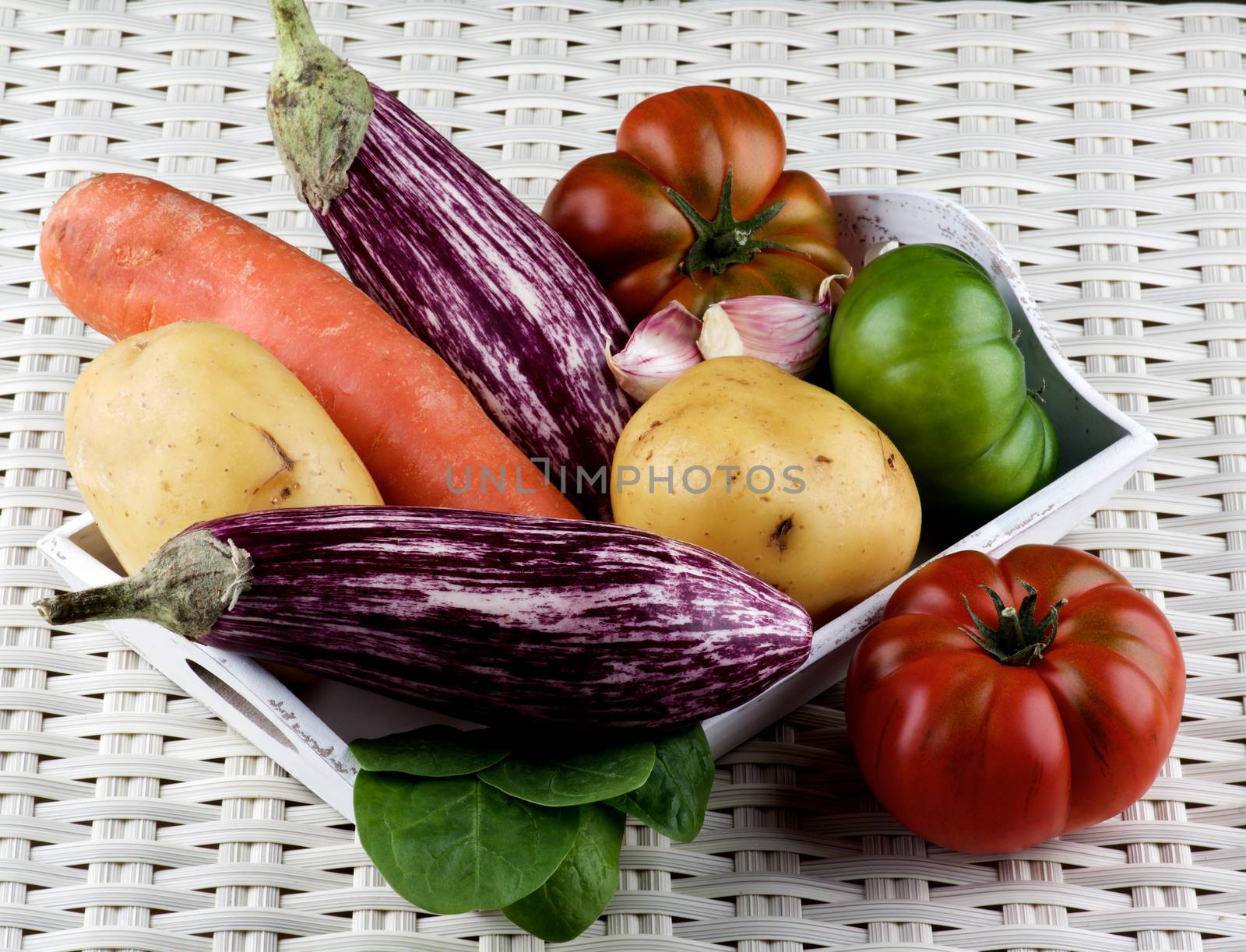 Arrangement of Fresh Raw Vegetables with Striped Eggplants, Potato, Carrot, Green and Red Tomatoes, and Garlic on Wooden Tray closeup on Wicker background