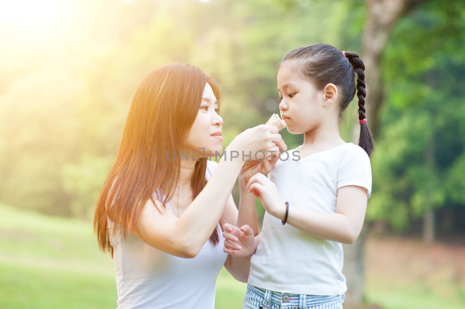 Lifestyle portrait of Asian mother and daughter eating sandwich in the park, Family outdoor fun, morning with sun flare.