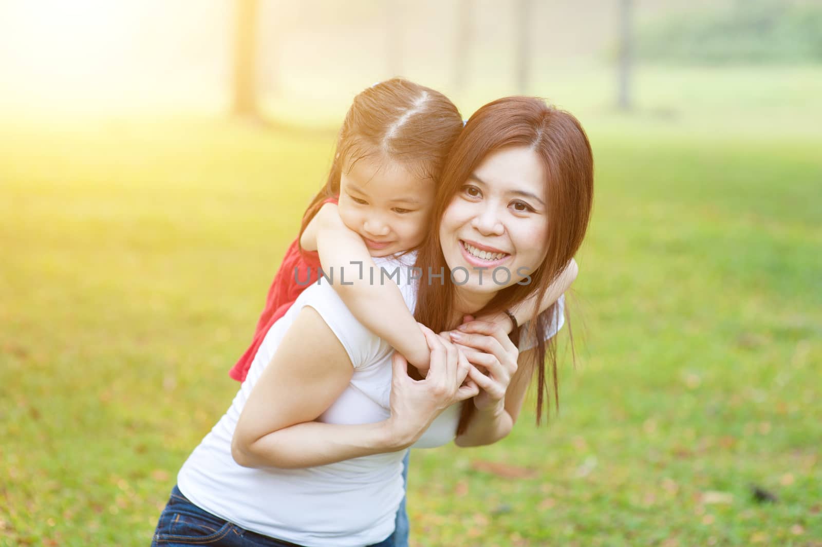 Pretty Asian woman piggyback her preschool daughter in back, little girl hugging her mother tenderly, morning with sun flare.