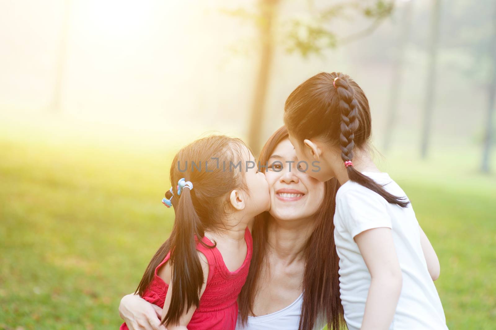 Lifestyle portrait of Asian mother and daughters kissing in the park, Family outdoor fun, morning with sun flare.