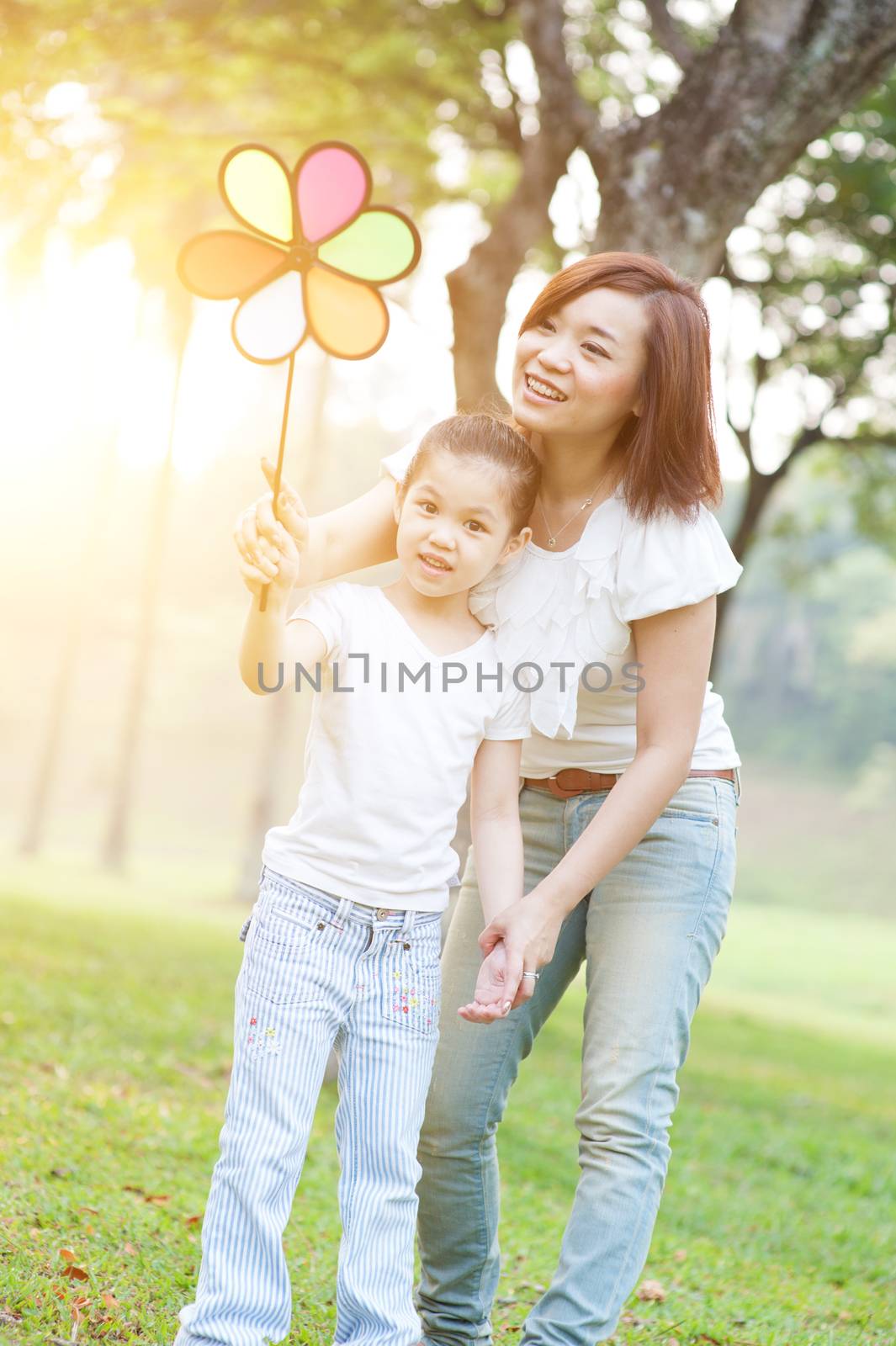 Happy Asian mother and daughter playing windmill in the park. Family outdoor fun, morning with sun flare.