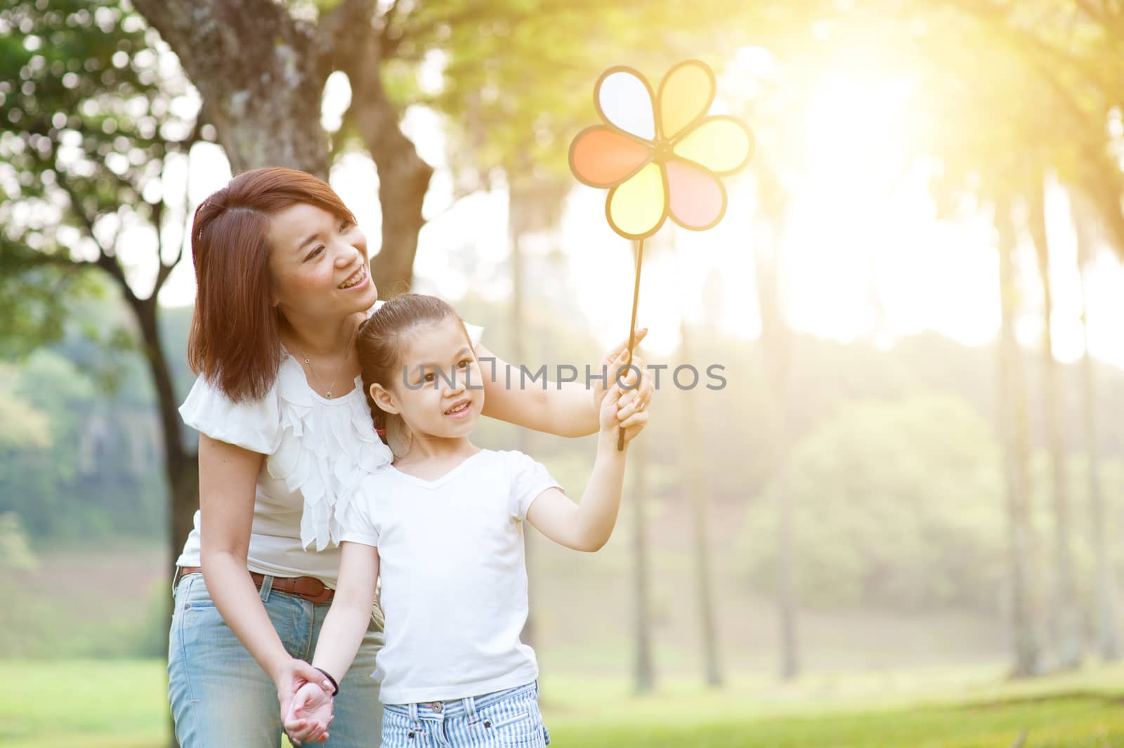 Happy Asian mother and daughter playing windmill toy in the park. Family outdoor fun, morning with sun flare.