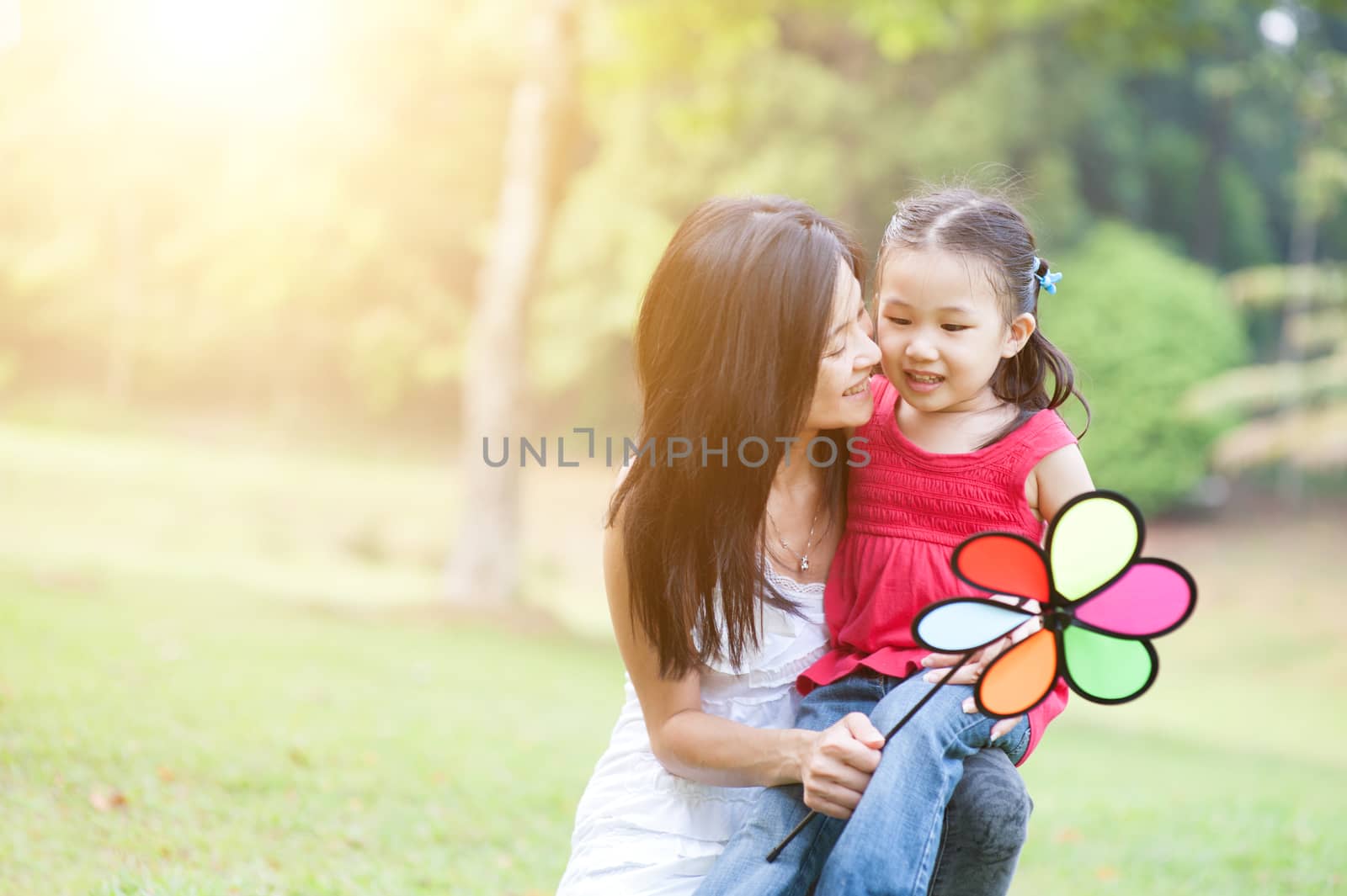 Lifestyle portrait of Asian mother and daughter playing windmill in the park, Family outdoor fun, morning with sun flare.