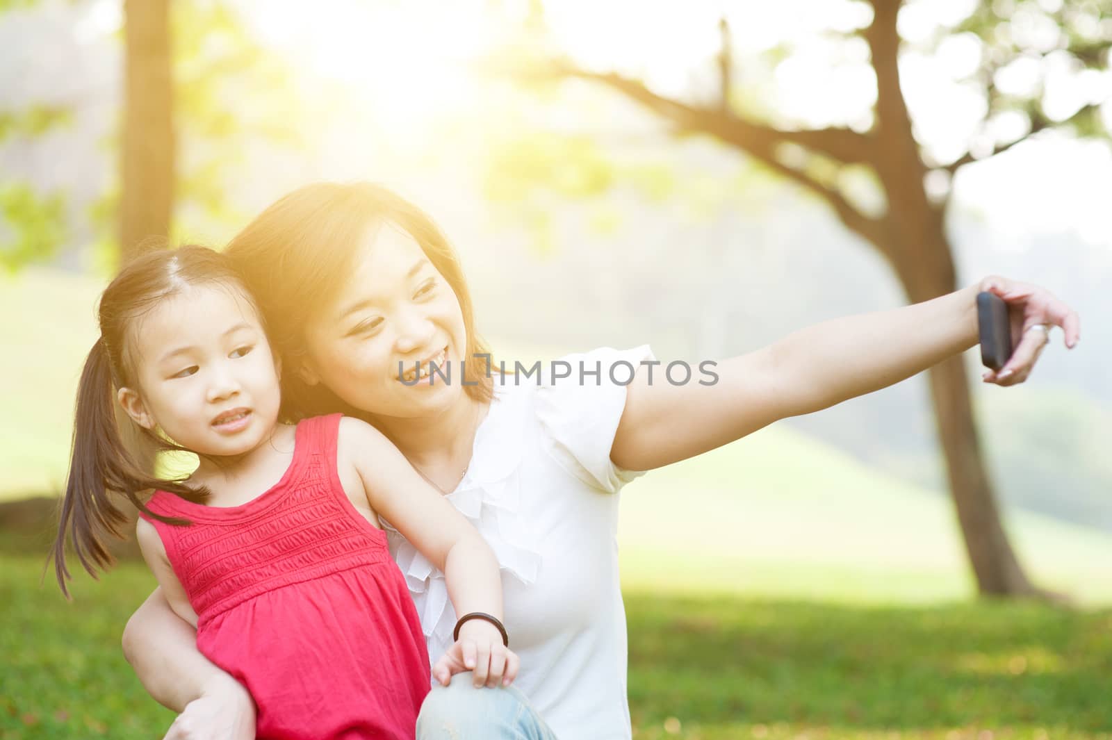 Lifestyle portrait of Asian mother and daughter taking selfie in happiness in the park, family outdoor fun, morning with sun flare.