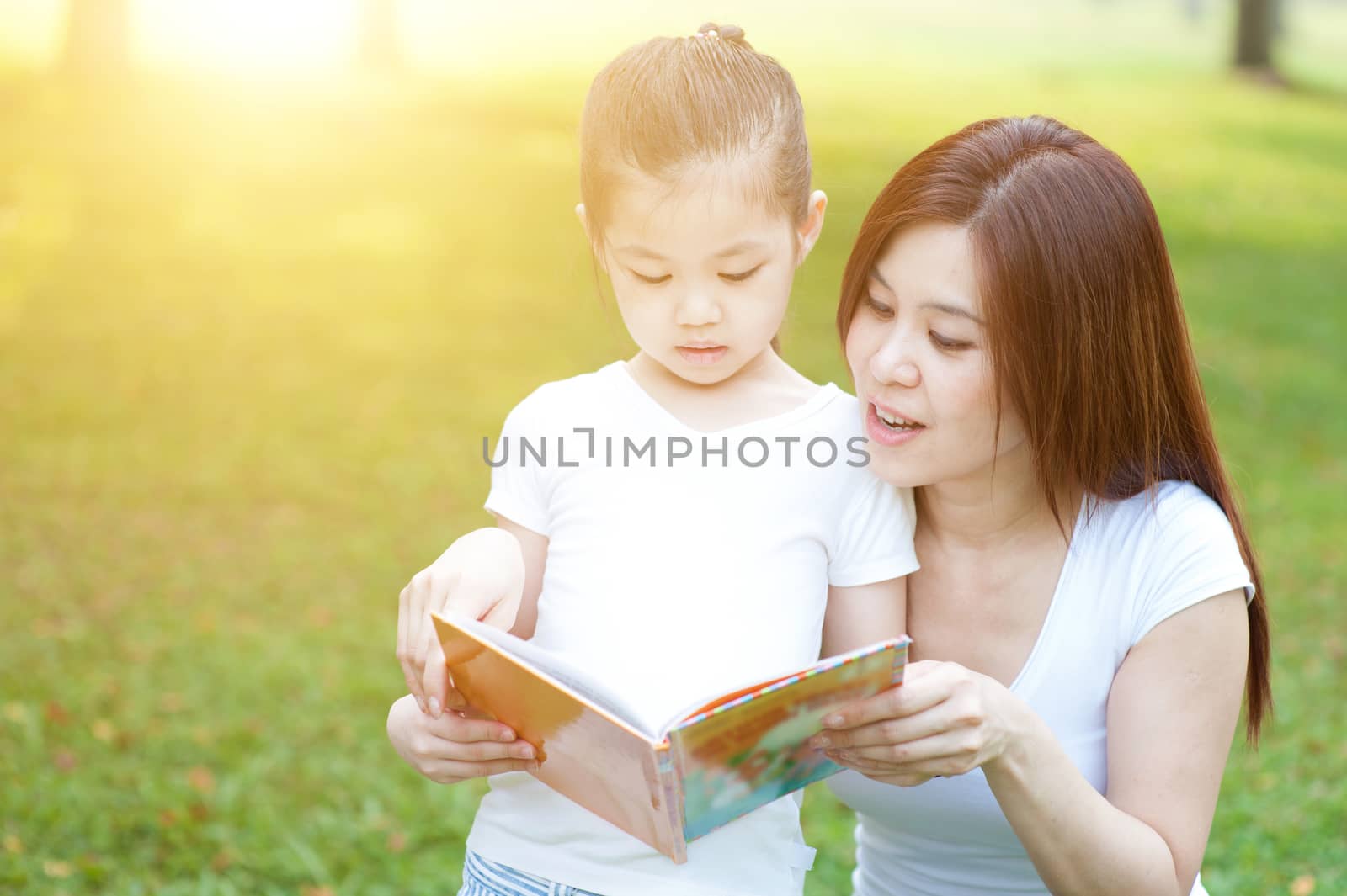 Asian mother and daughter are reading book and having fun in nature. Family outdoor fun, morning with sun flare.