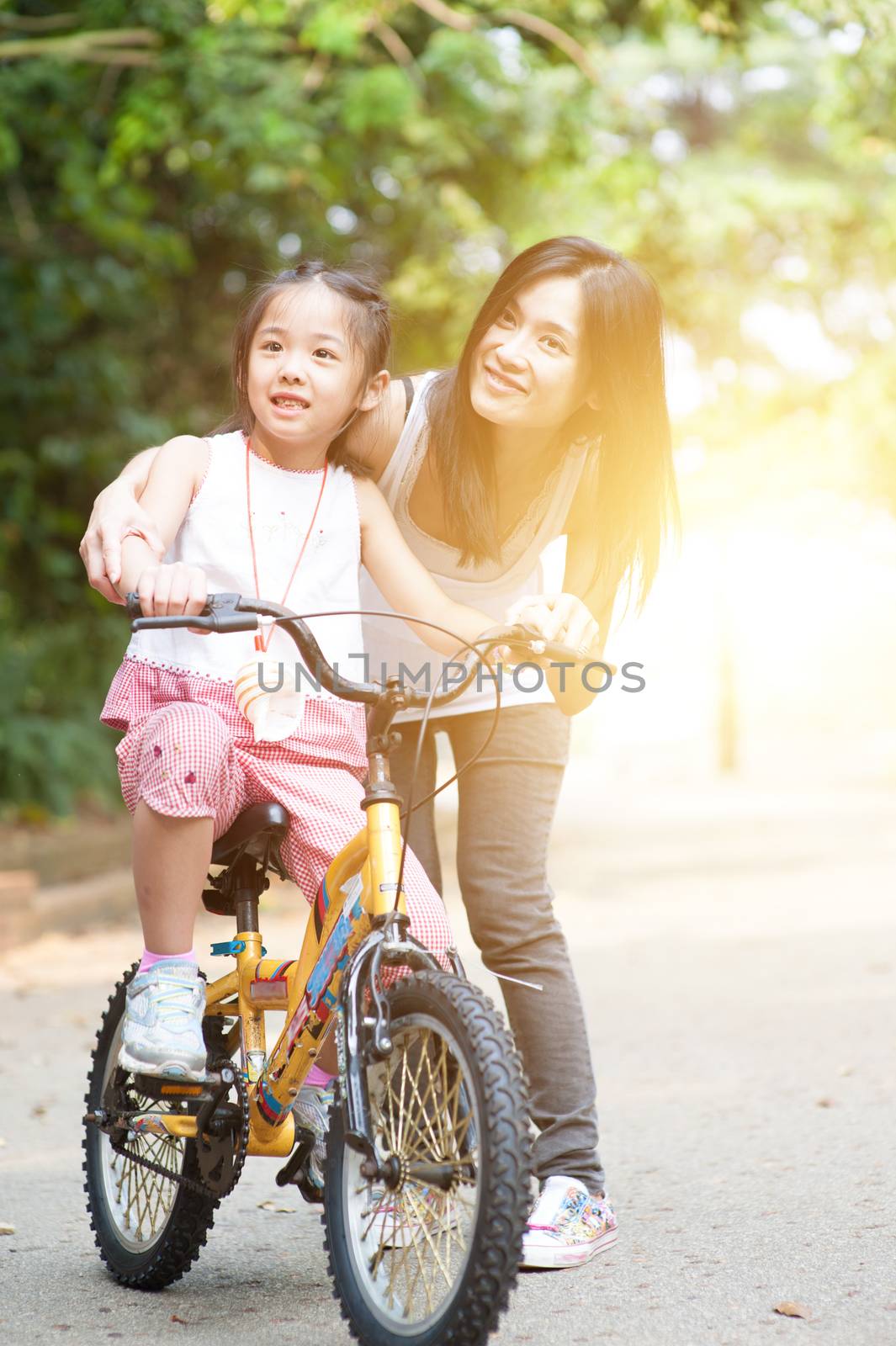 Asian little girl learning ride on bicycle with help of mother in the park, family outdoor fun activity.