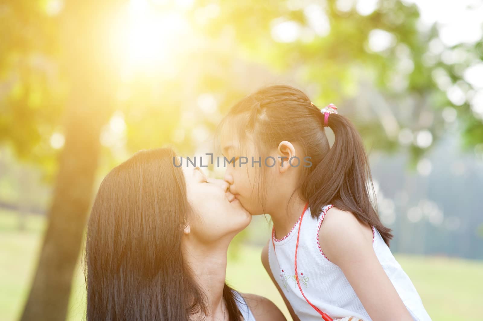 Lifestyle portrait mom and daughter kissing in happiness at the outside in the park. Family outdoor fun, morning with sun flare.