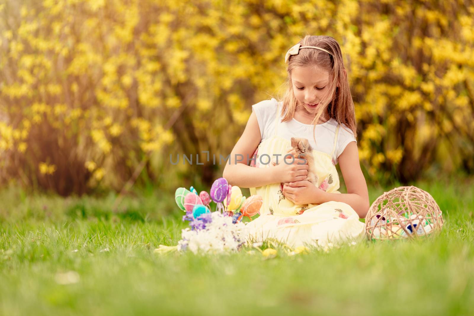 Beautiful smiling little girl holding cute bunny and sitting on the grass with Easter eggs in spring holidays. Copy space.