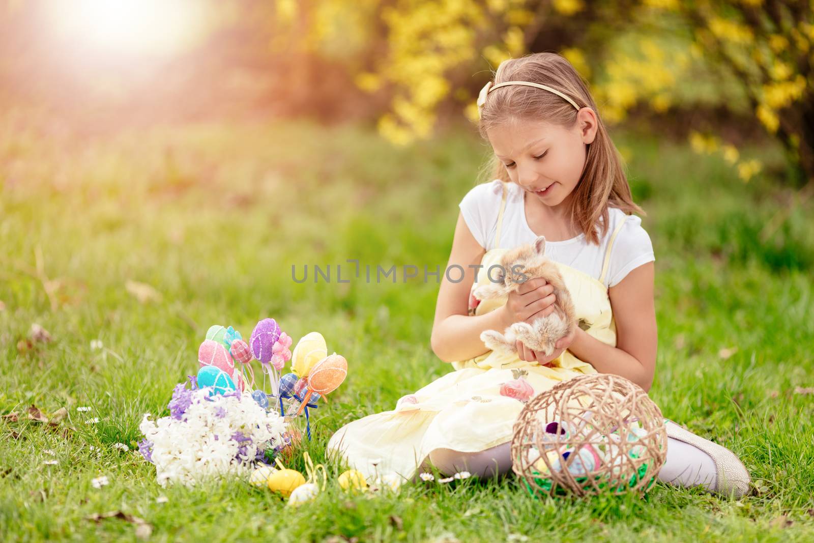 Beautiful smiling little girl holding cute bunny and sitting on the grass with Easter eggs in spring holidays. 