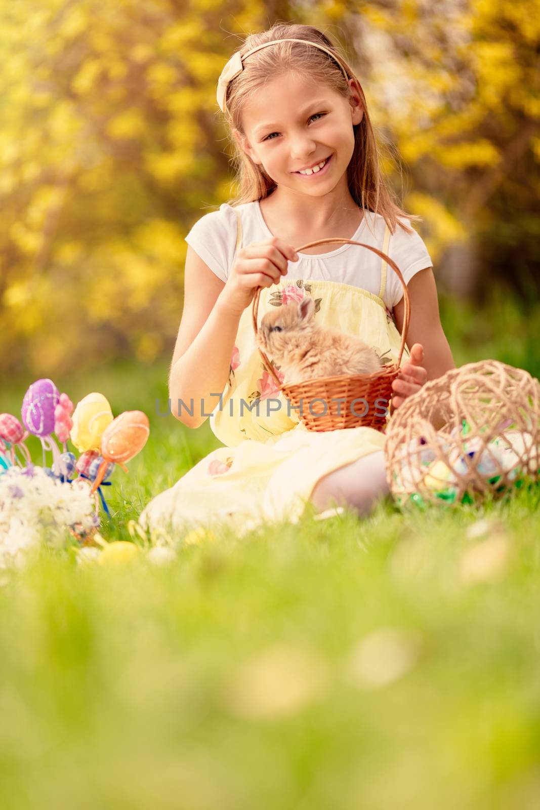 Beautiful smiling little girl holding cute bunny in the basket and sitting on the grass with Easter eggs in spring holidays. Looking at camera. Copy space.