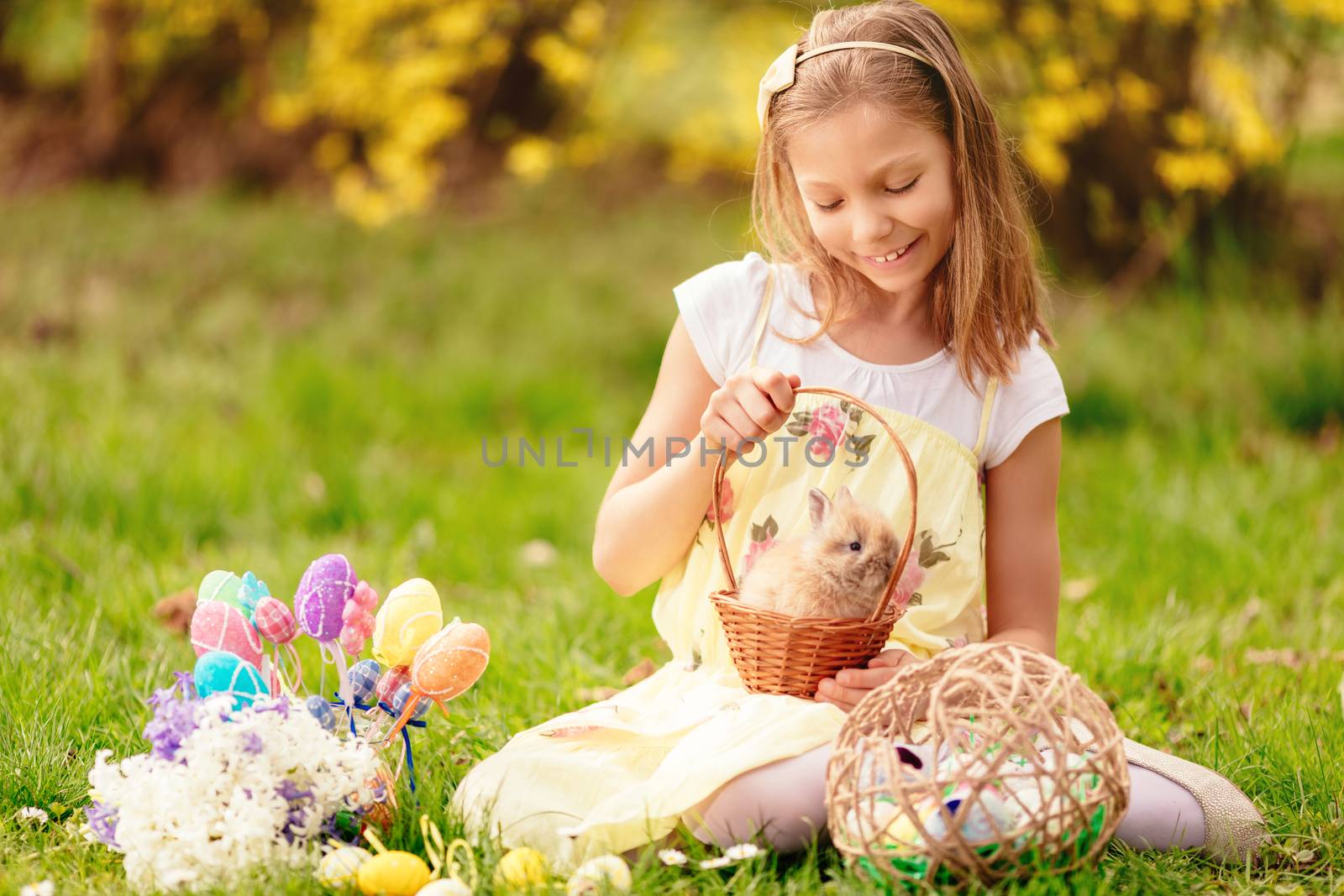 Beautiful smiling little girl holding cute bunny in the basket and sitting on the grass with Easter eggs in spring holidays. 