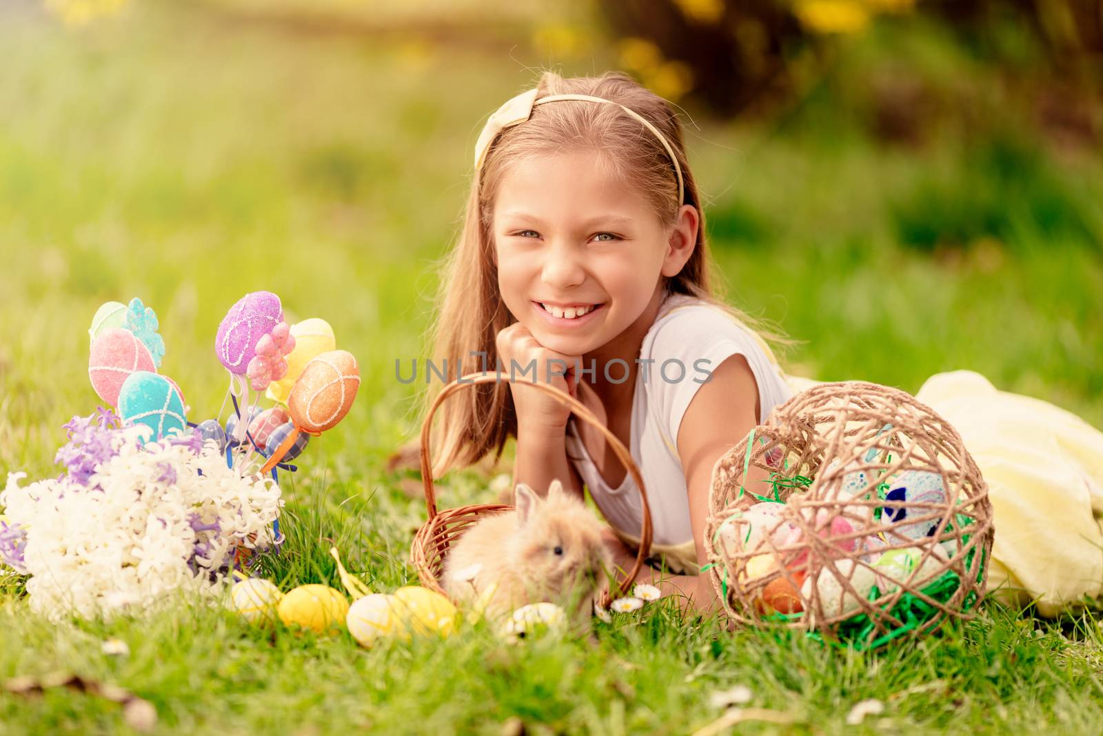 Beautiful smiling little girl with cute bunny in the basket and Easter eggs on the grass in spring holidays. Looking at camera.