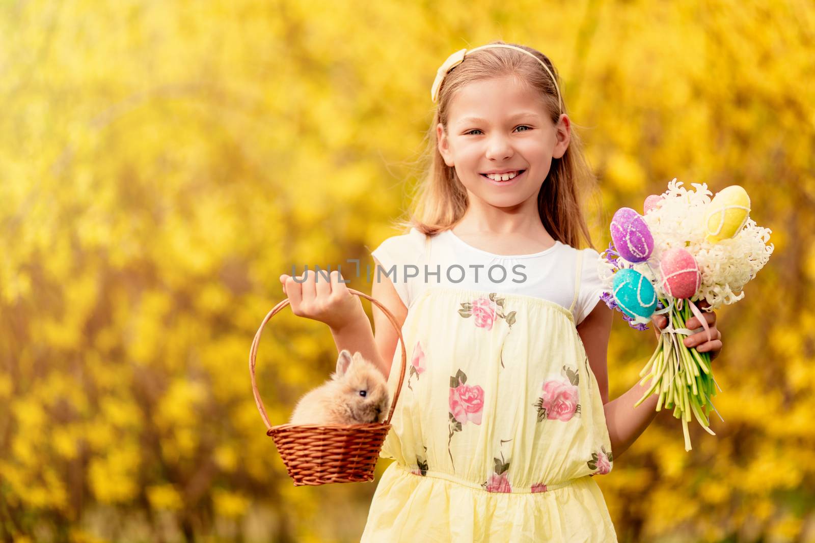 Beautiful smiling little girl holding cute bunny in the basket and bouquet white flowers and Easter eggs in spring holidays. Looking at camera.