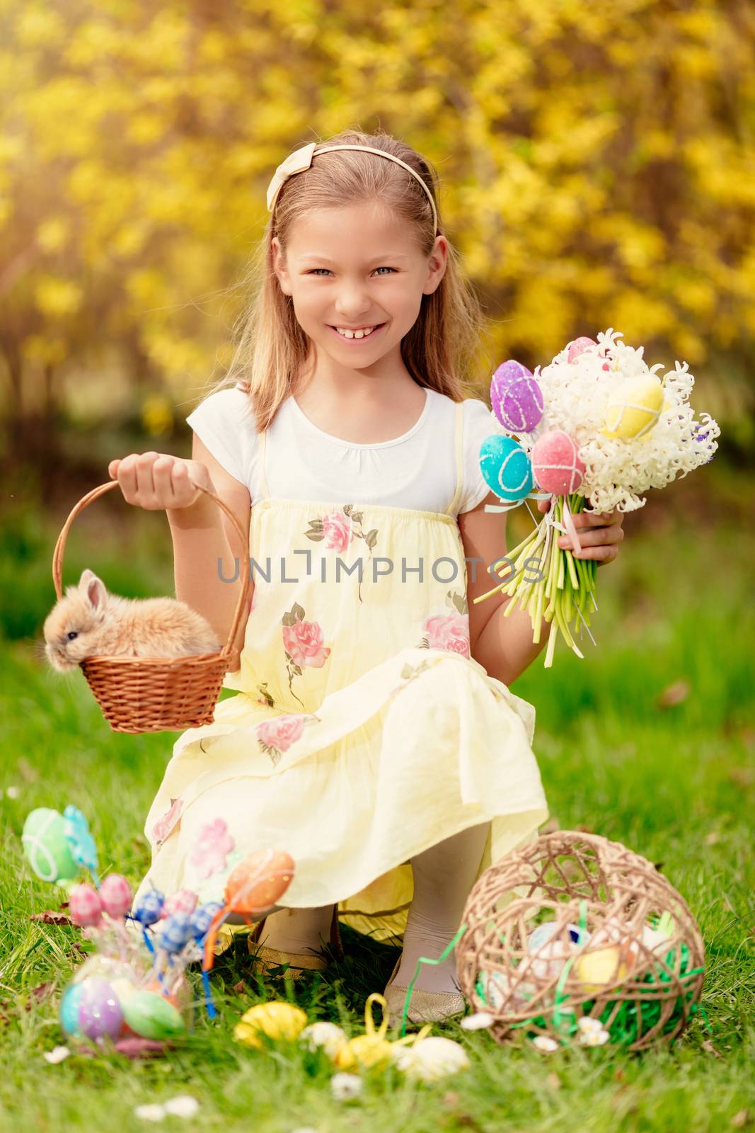 Beautiful smiling little girl holding cute bunny in the basket and bouquet white flowers and Easter eggs in spring holidays. Looking at camera.