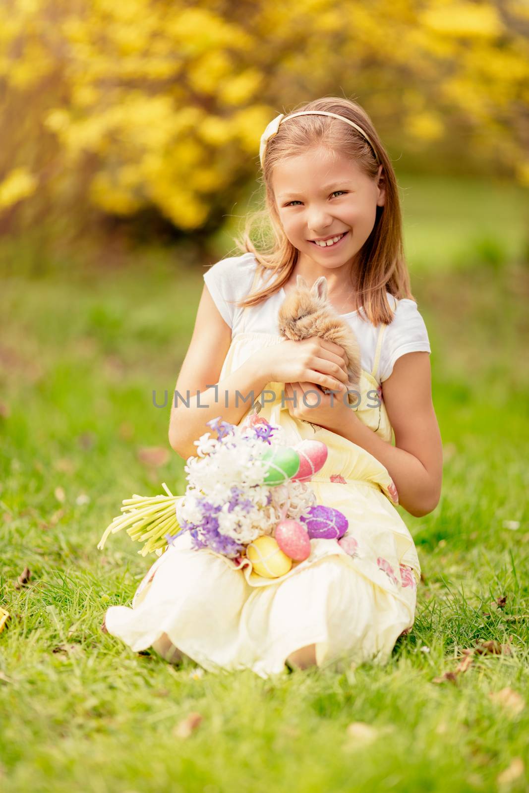 Beautiful smiling little girl holding cute bunny and bouquet white flowers and Easter eggs in spring holidays.