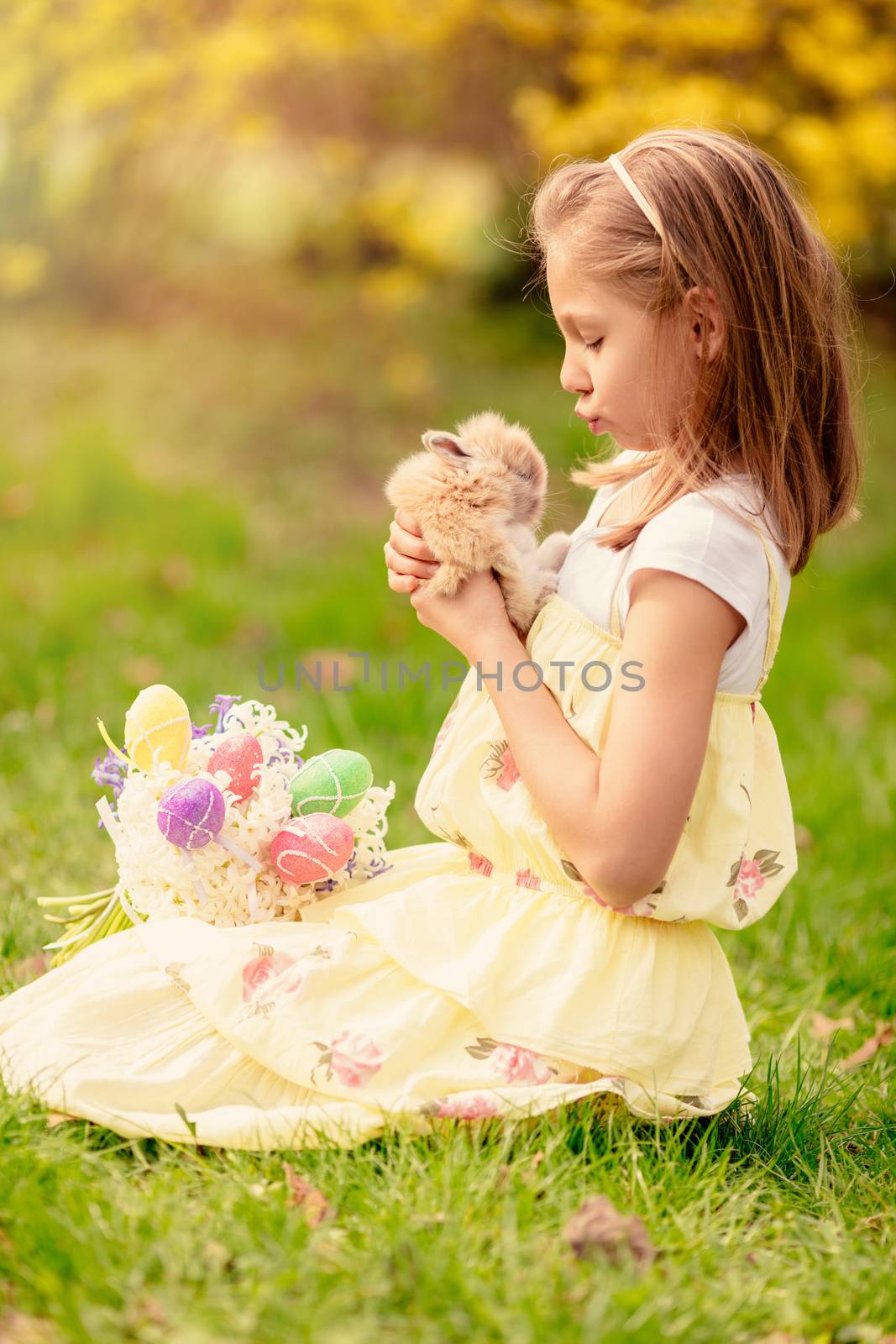 Beautiful smiling little girl holding cute bunny and bouquet white flowers and Easter eggs in spring holidays.