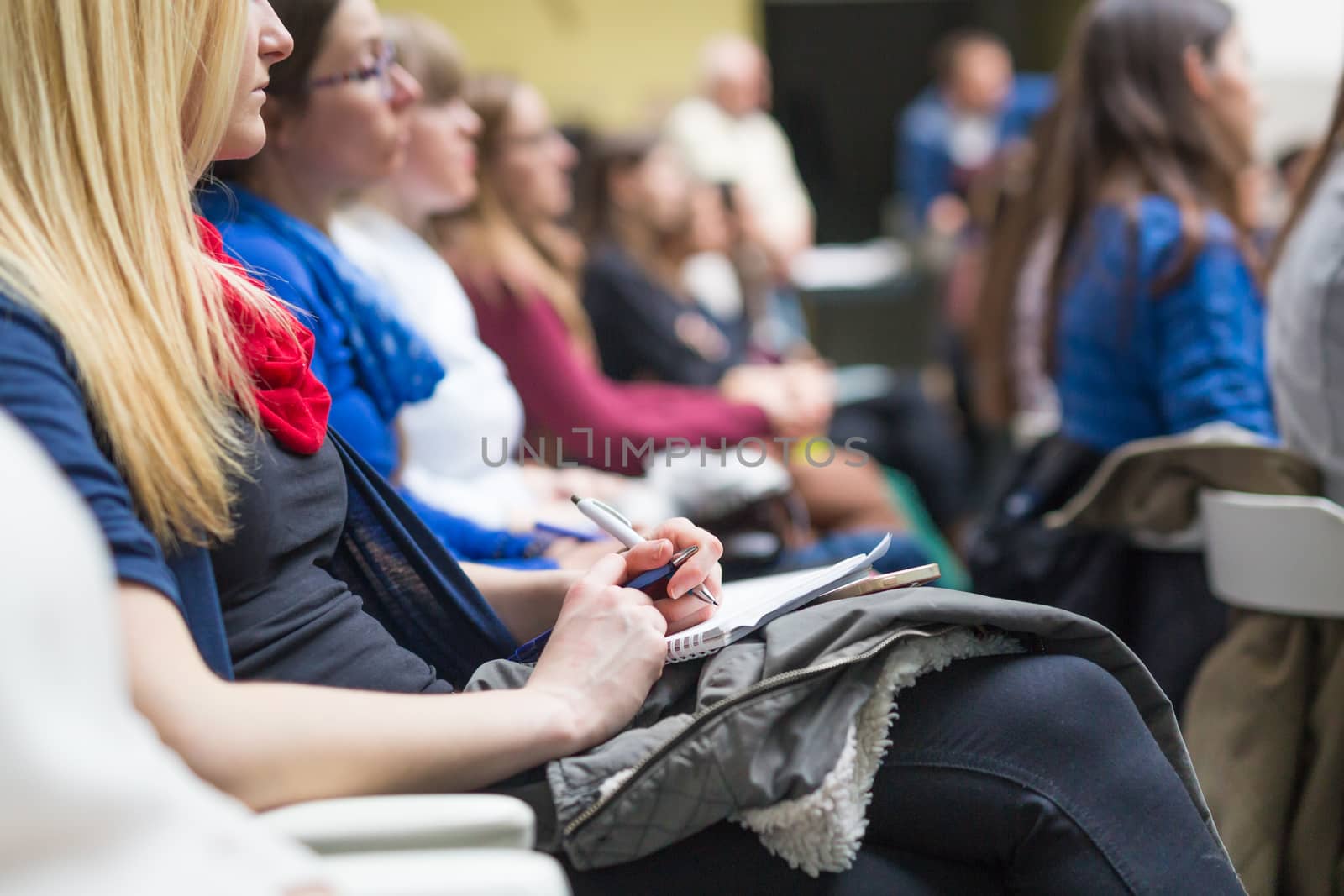 Female hands holding pens, notebook and phone and making notes at conference lecture. Event participants in conference hall.