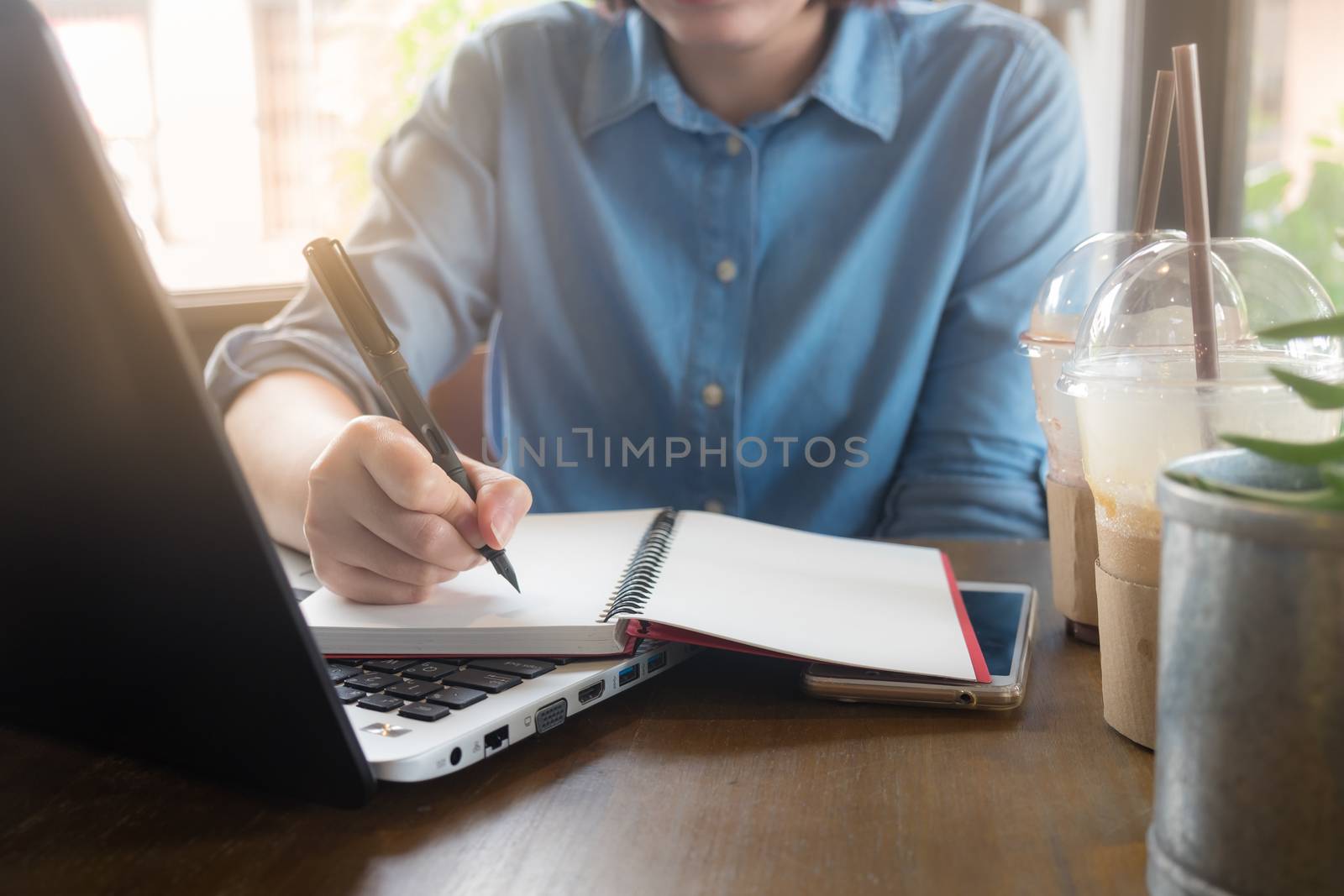 Young Asian hipster woman writing on notebook by right hand while working with laptop computer in coffee shop. Freelance business starup activity concept