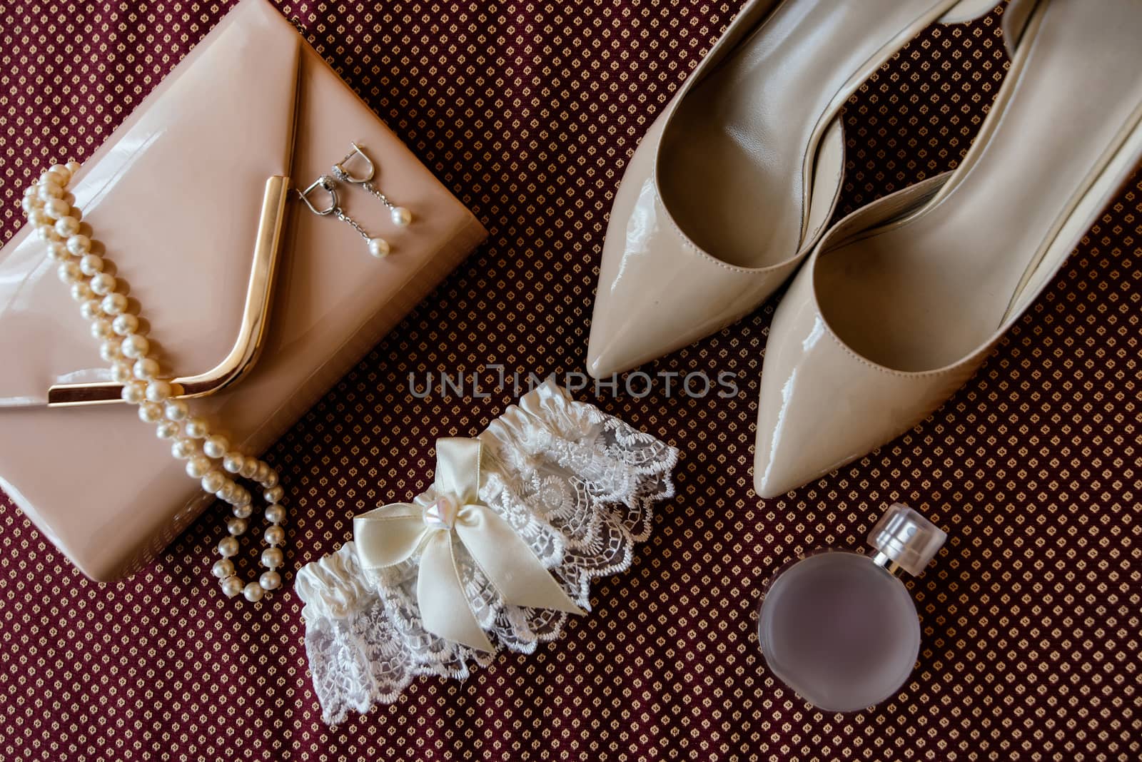 Bride's shoes, garter, perfume, necklace and clutch on burgundy cloth surface by d_duda