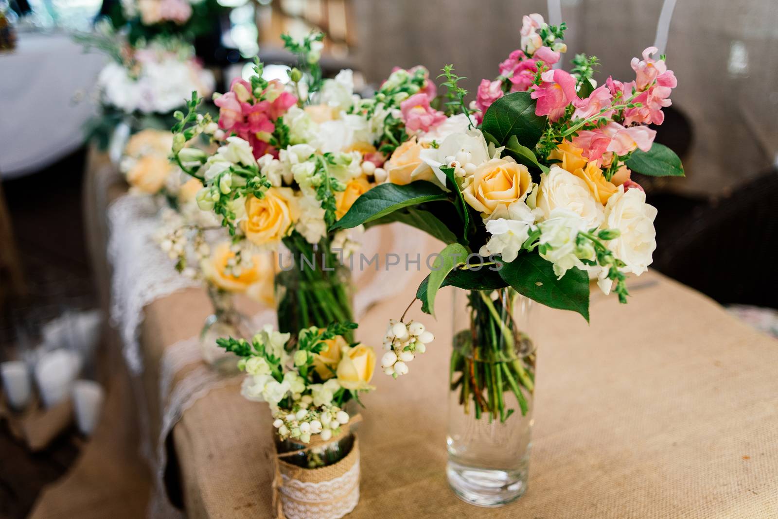 Wedding bouquets of yellow, white and pink roses are in vases on dinner table