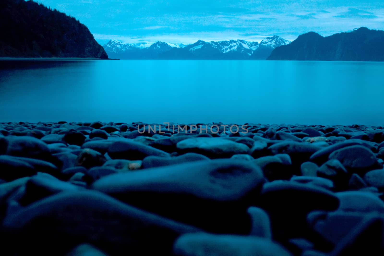 Smooth beach stones on the shore in the foreground of a landscape photo overlooking the ocean and mountains in Seward Alaska at midnight. 