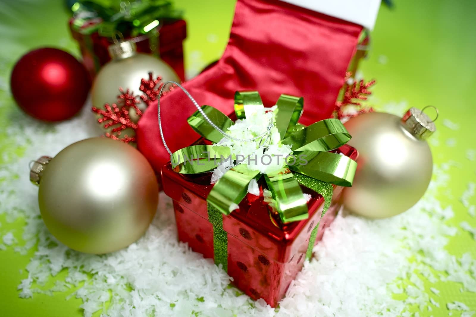 Holiday Present - Red Present Box with Green Glossy Bow. Some Christmas Ornaments and Fake Snow. Green Background