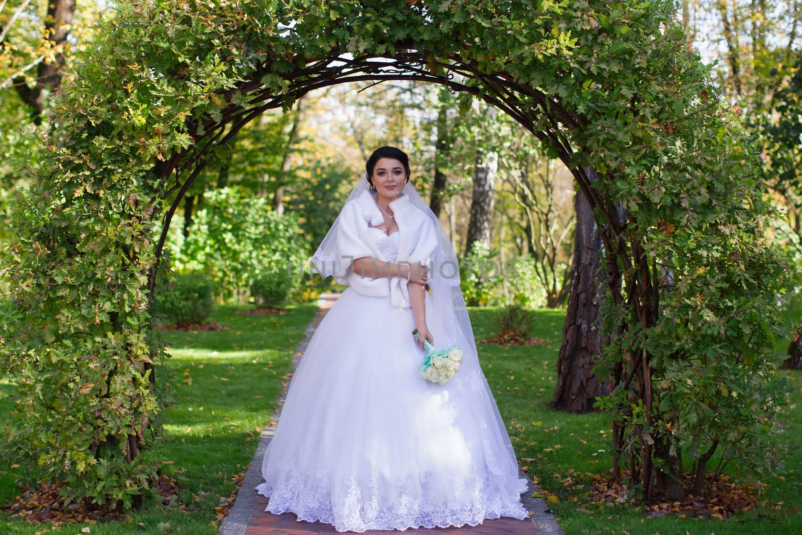 A stylish bride with a bouquet in her hands is standing under a green arch