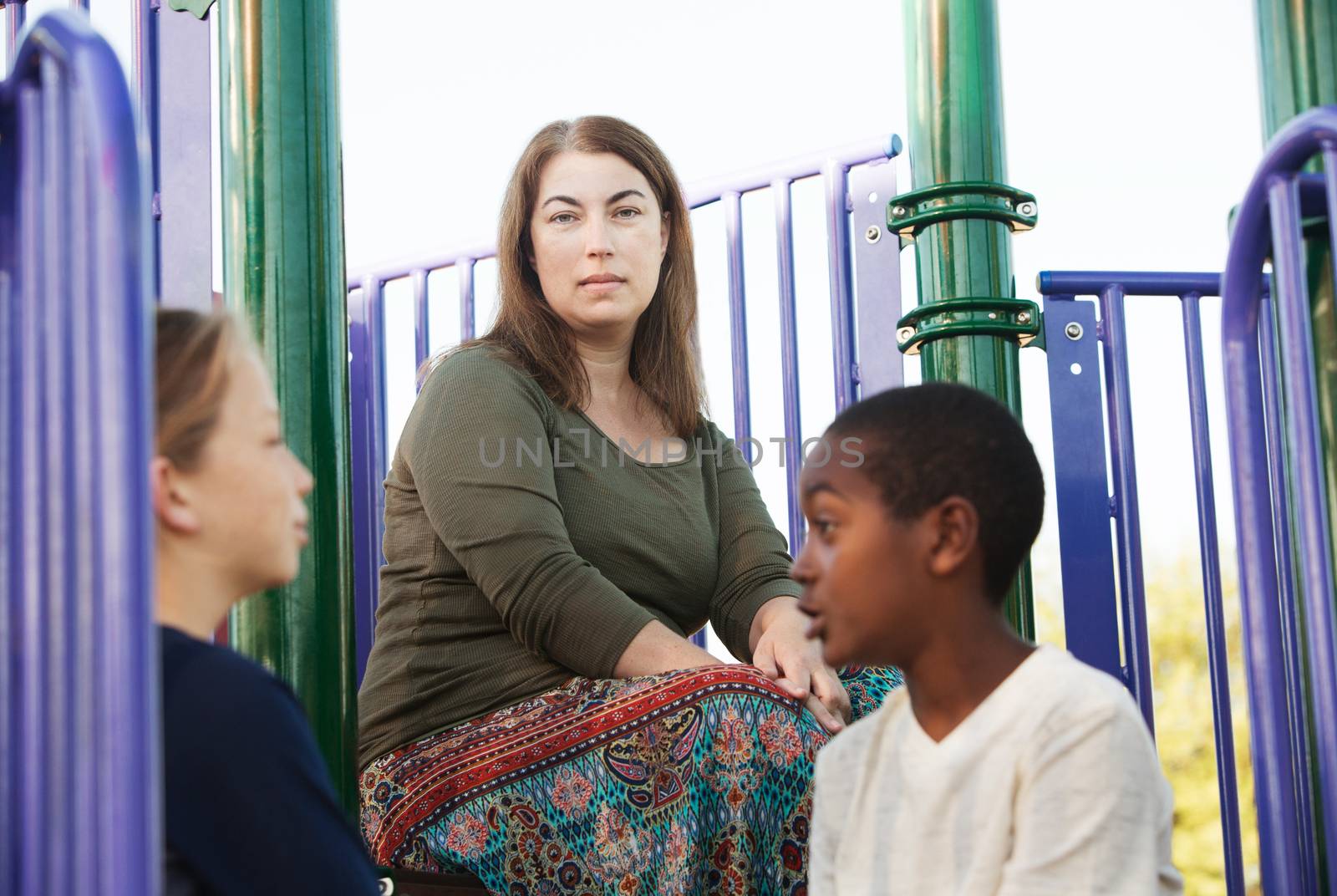 Two children talking to each other on playground set with mother watching from behind them