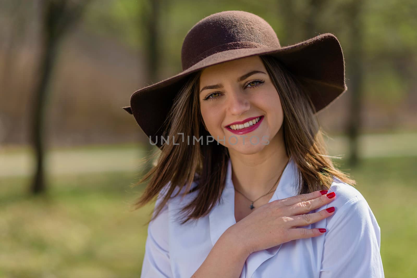 Portrait of a smiling young woman, holding her hand on her shoulder and wearing a hat in a park during spring. Woman bowed her head head to the left side of frame. Medium shot. Shallow depth of field.